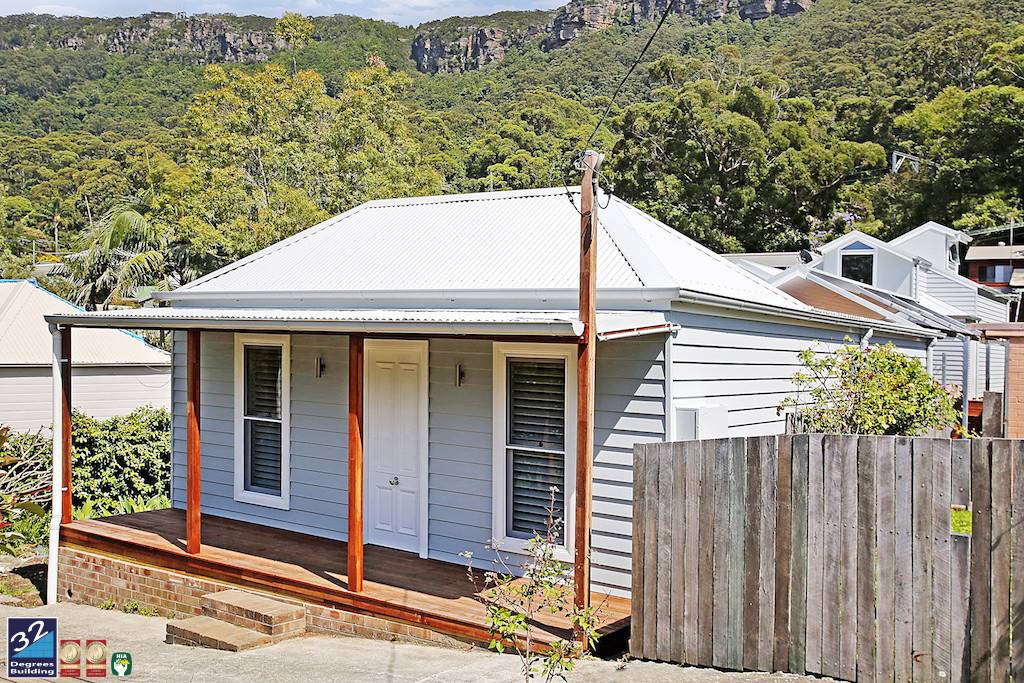 A small, light blue house with a white roof and front porch stands beside a wooden fence, surrounded by trees and hills in the background.
