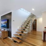 stairs-first-floor-addition-bossley-park Modern living space with wooden floors, a floating wooden staircase with glass railing, a TV mounted on a black accent wall, and a sideboard. The room is bright, with ample natural and artificial light.