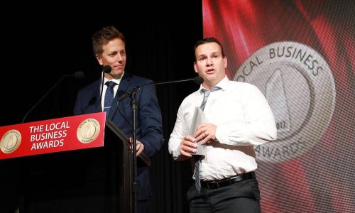 business_man_of_the_year_2017 Two men stand on stage at a podium during The Local Business Awards. One in a blue suit speaks into a microphone, while the other, wearing a white shirt and tie, holds a trophy. A large awards logo is displayed in the background.