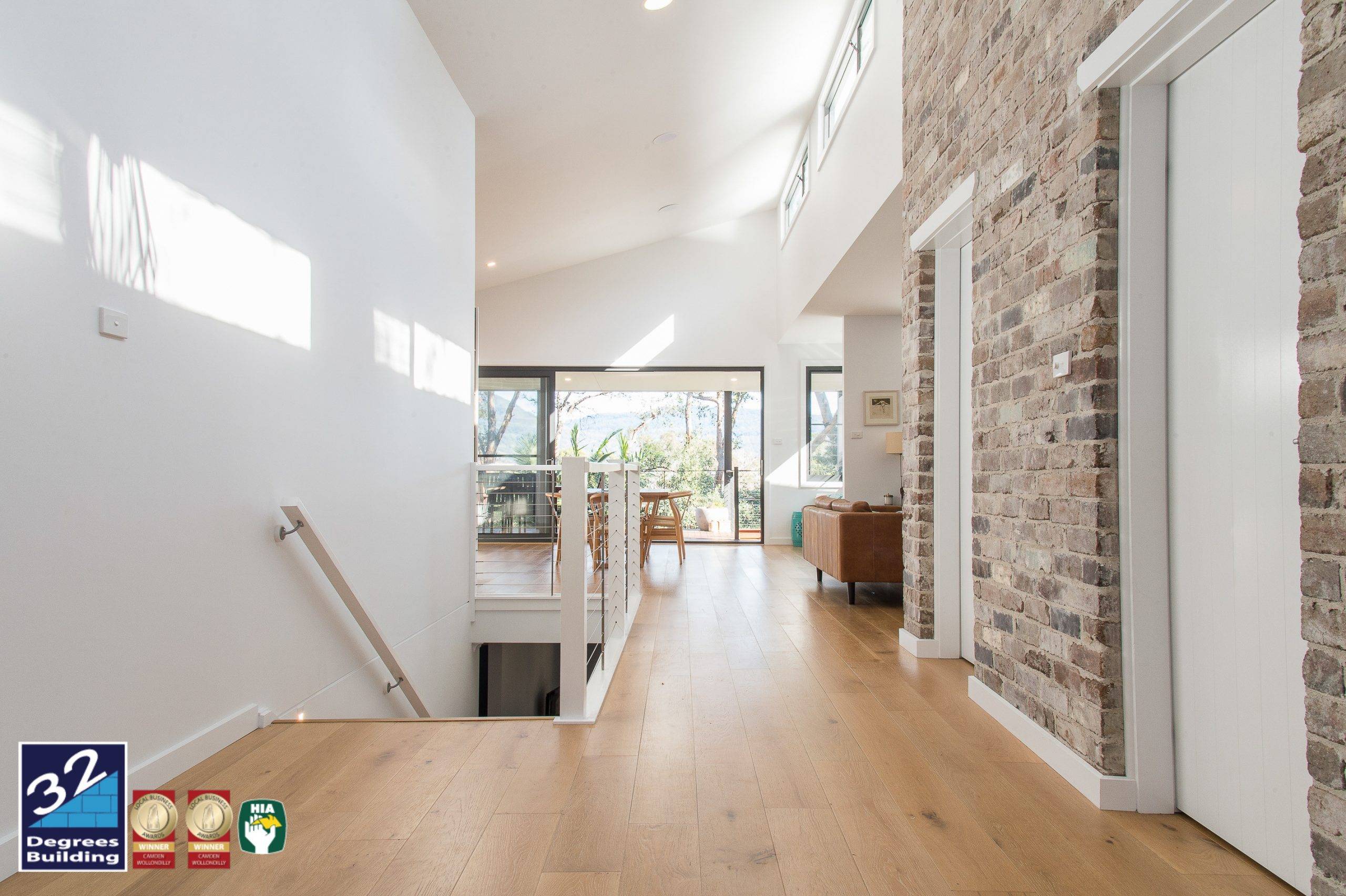 Bright modern hallway with wooden flooring, exposed brick walls, and large windows letting in natural light. A staircase leads down, and a dining area and living room are visible toward the back. Building awards are displayed at the bottom left.