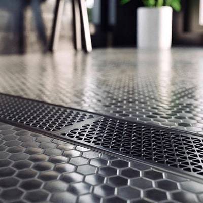 Close-up of a metallic floor with a hexagonal tile pattern and a rectangular grid drain in the center; a white plant pot and furniture are blurred in the background.