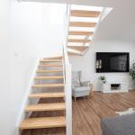 second storey addition prospect (15) Modern, bright living room with wooden open stairs, white walls, wooden floor, a gray armchair, a TV on the wall, and a small white cabinet with plants. Natural light fills the space.