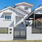 A modern white two-story house with a front porch, grey steps, a black metal gate, a carport on the right, and a small front lawn under a clear blue sky. There are award icons and a logo in the bottom left corner.