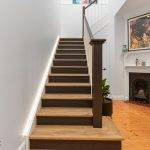 First Floor Addition Earlwood-10 A straight wooden staircase with dark railings leads upward in a modern home with white walls, a fireplace, artwork, and hardwood floors.
