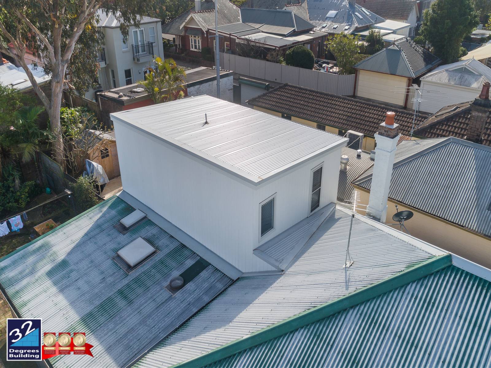 Aerial view of a white, modern rooftop extension added to a house with a metal roof, surrounded by neighboring homes and trees in a suburban area. 32 Degrees Building logo is visible in the bottom left corner.
