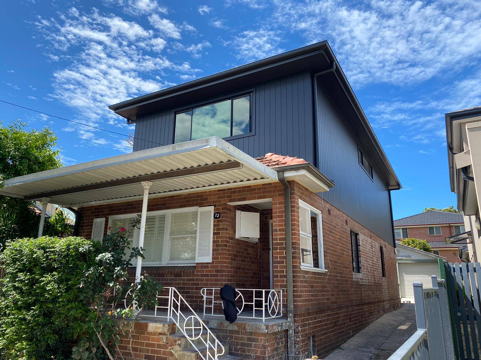 A two-story house with a modern dark gray upper level and a brick lower level, featuring a metal awning over the porch and a small garden in front, under a bright blue sky with scattered clouds.