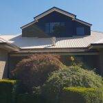A modern house with a light-colored metal roof, a dark upper dormer with large windows, brick walls, and well-maintained green shrubs and bushes in the front garden under a clear blue sky.