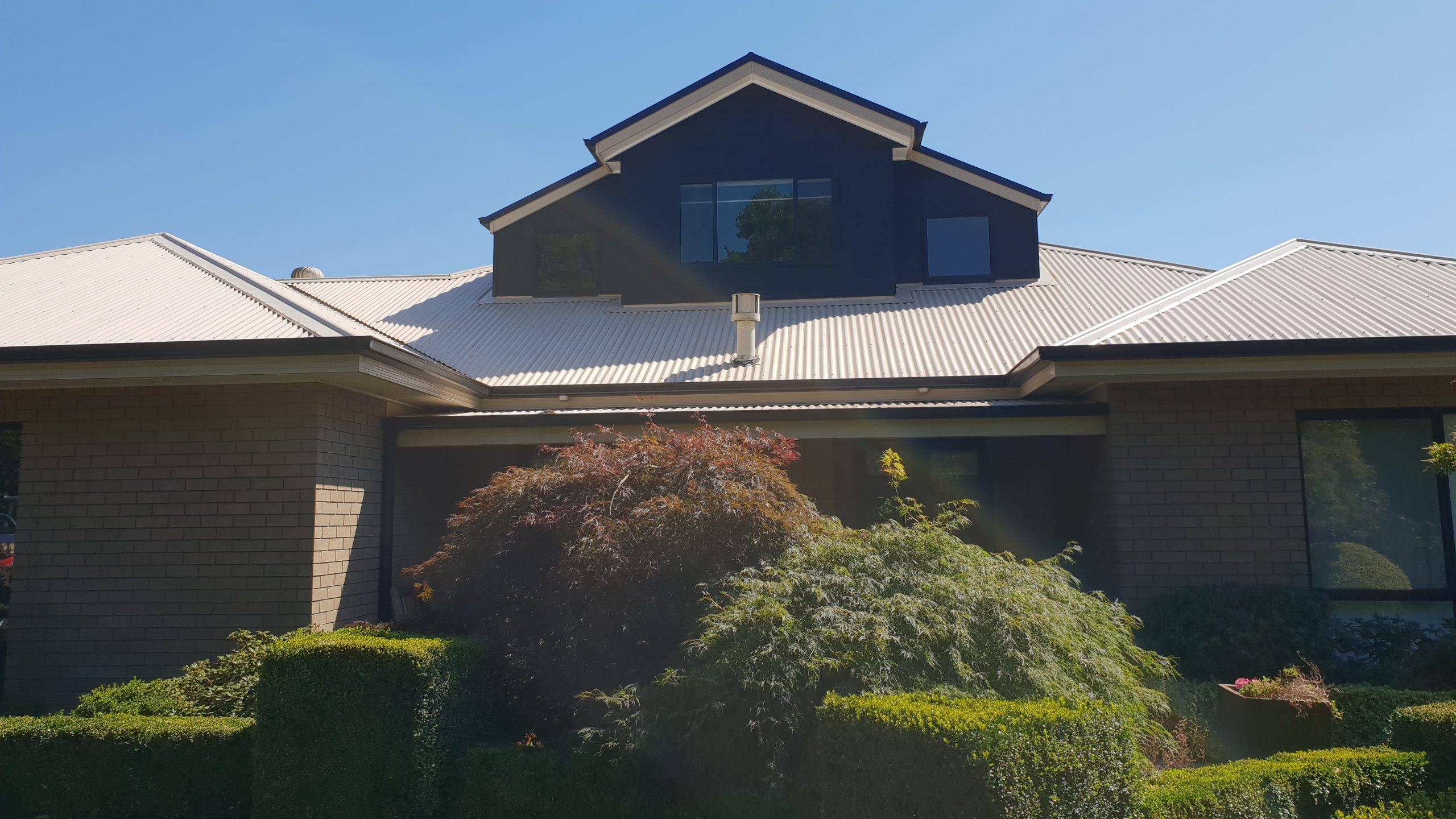 A modern house with a light-colored metal roof, a dark upper dormer with large windows, brick walls, and well-maintained green shrubs and bushes in the front garden under a clear blue sky.