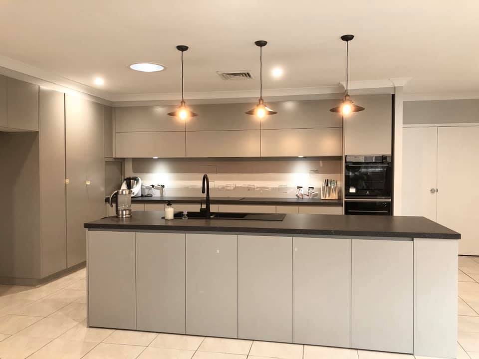 Modern kitchen with light gray cabinets, black countertops, and a central island. Three pendant lights hang above the island. Appliances, kettle, and utensils are visible on the back counter. The floor is tiled in white.