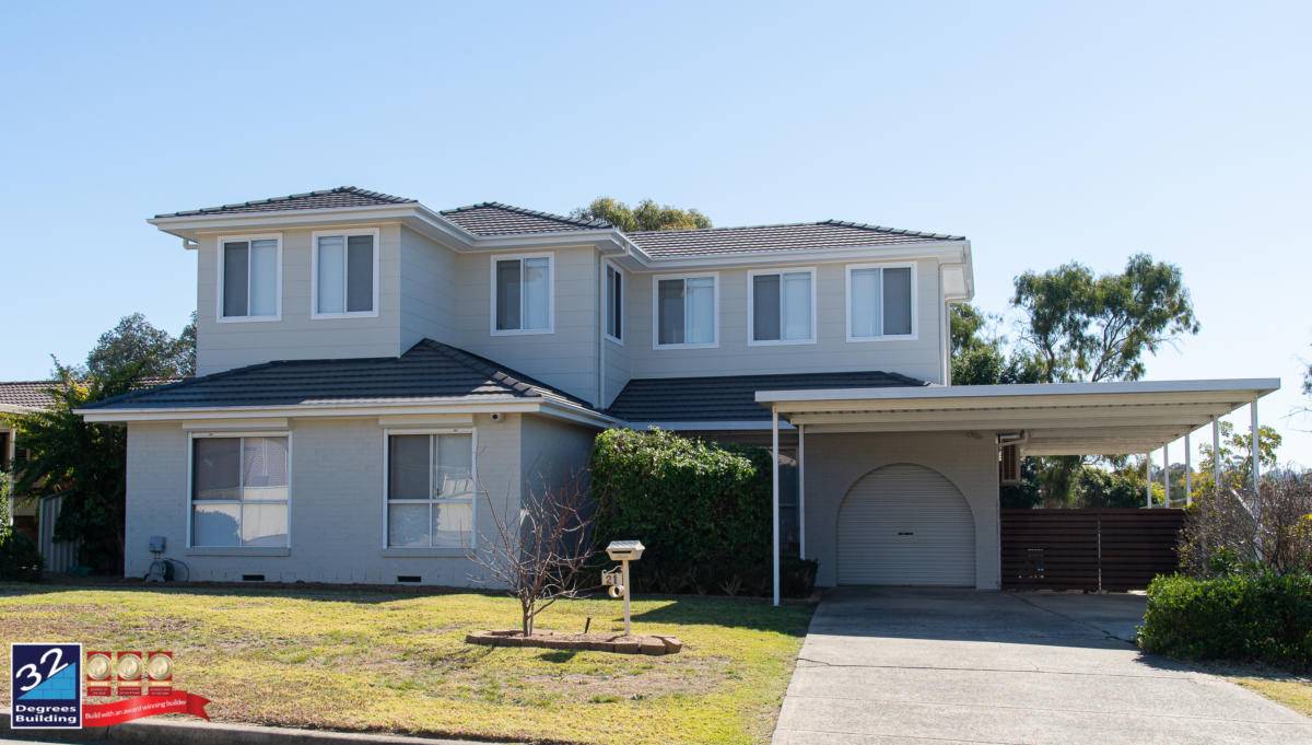 A two-story suburban house with light gray exterior, large windows, a driveway leading to a carport, a garage, and a small front yard with grass and a mailbox. A 32 Degrees Building logo is in the lower left corner.