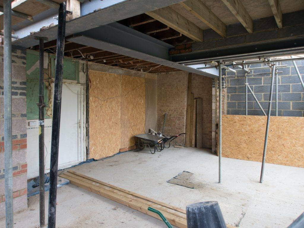 An unfinished room under construction with exposed beams, a wheelbarrow, plywood sheets, and various construction materials scattered across the floor. The walls are partially covered with insulation and bricks.