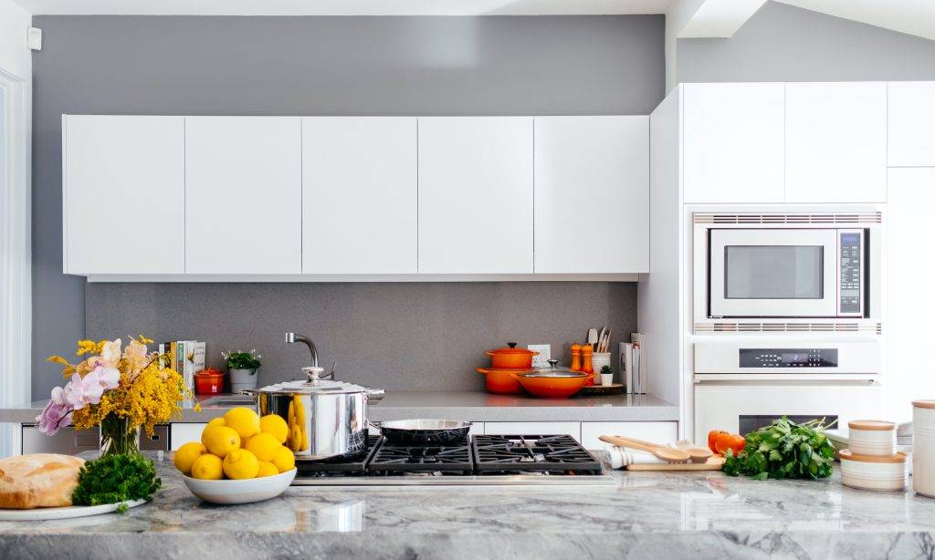 Modern kitchen with white cabinets, a built-in microwave, and a gray backsplash. The marble counter holds lemons, herbs, bread, a pot, flowers, and various kitchenware in bright orange and silver.