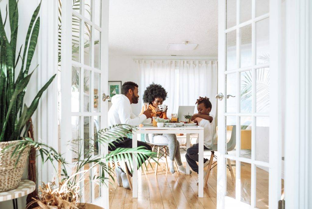 A family of three, two adults and one child, sits around a dining table eating breakfast in a bright, sunlit room with large windows and green plants. The atmosphere appears warm and cozy.