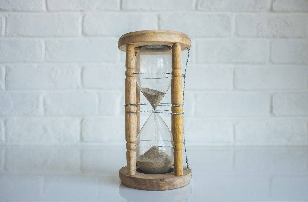A wooden hourglass with sand flowing from the upper to the lower chamber sits on a reflective white surface in front of a white brick wall.