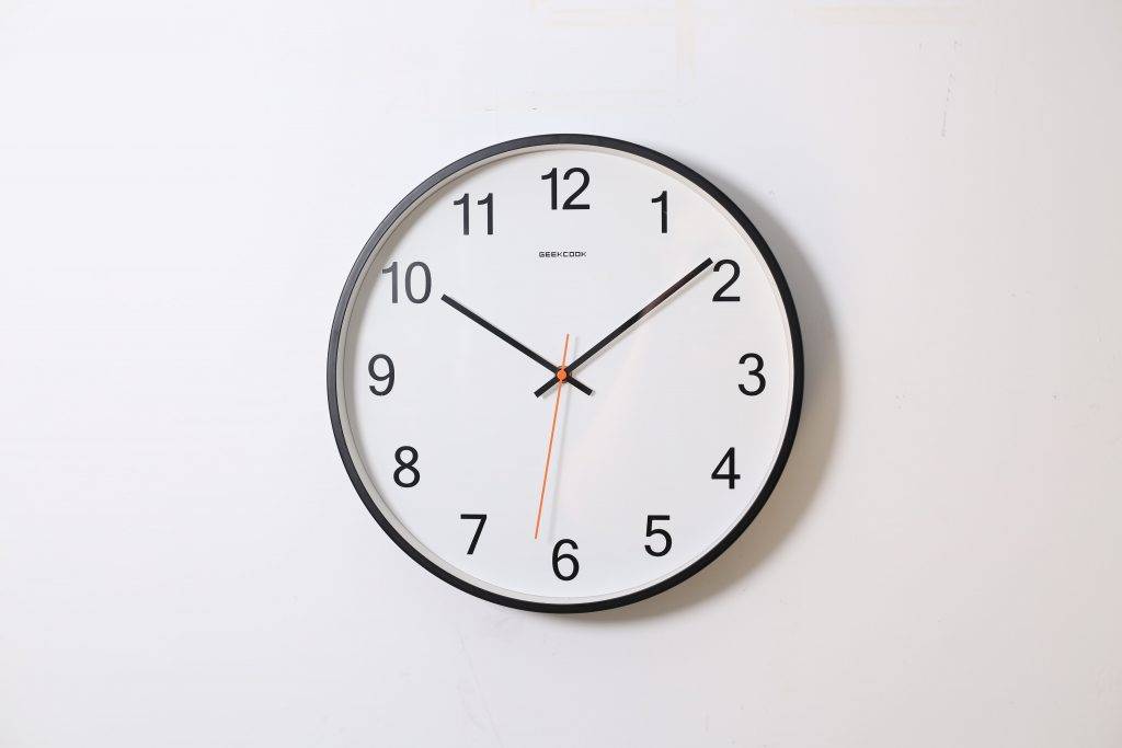 A round wall clock with a white face, black frame, black hour and minute hands, and a thin orange second hand, showing the time as 10:10:31 against a plain white wall.