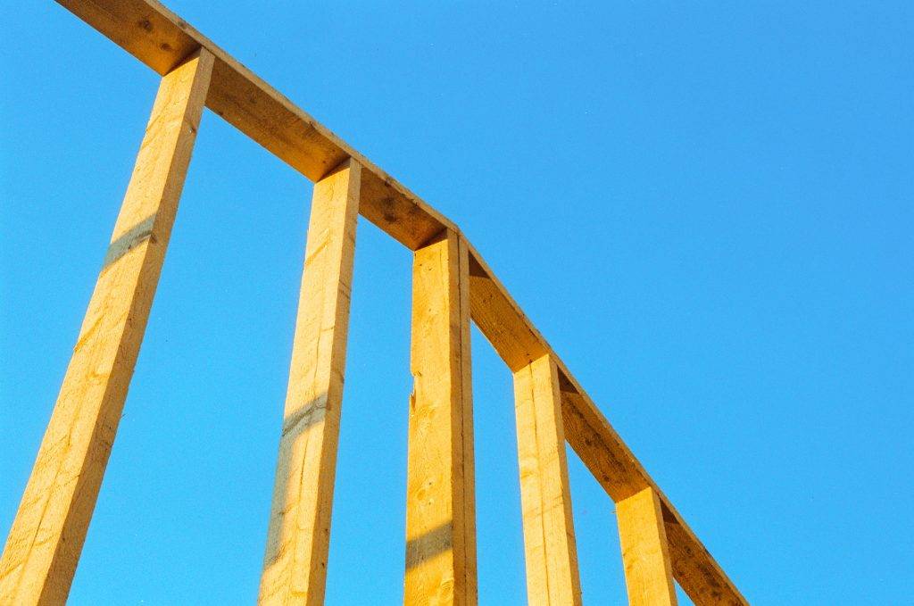Wooden framing beams of a building under construction are shown at an angle against a clear blue sky.