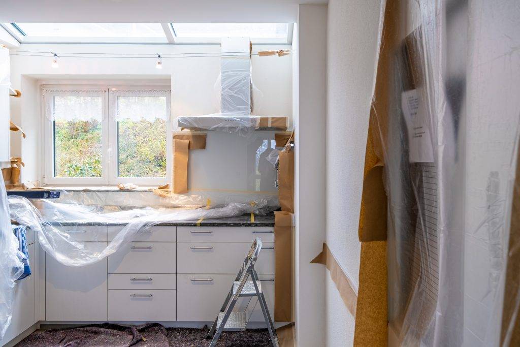 A modern kitchen under renovation, with cabinets and appliances covered in plastic sheets and cardboard. A step ladder stands in front of the countertop, and sunlight streams in through the window and skylight above.