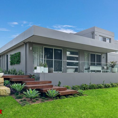 Ground Floor Extension - Grasmere--5 A modern, two-story gray house with large windows, a wooden deck, outdoor seating, and landscaped garden featuring succulents and a green lawn. A 32 Degrees Building logo with award icons is in the corner.