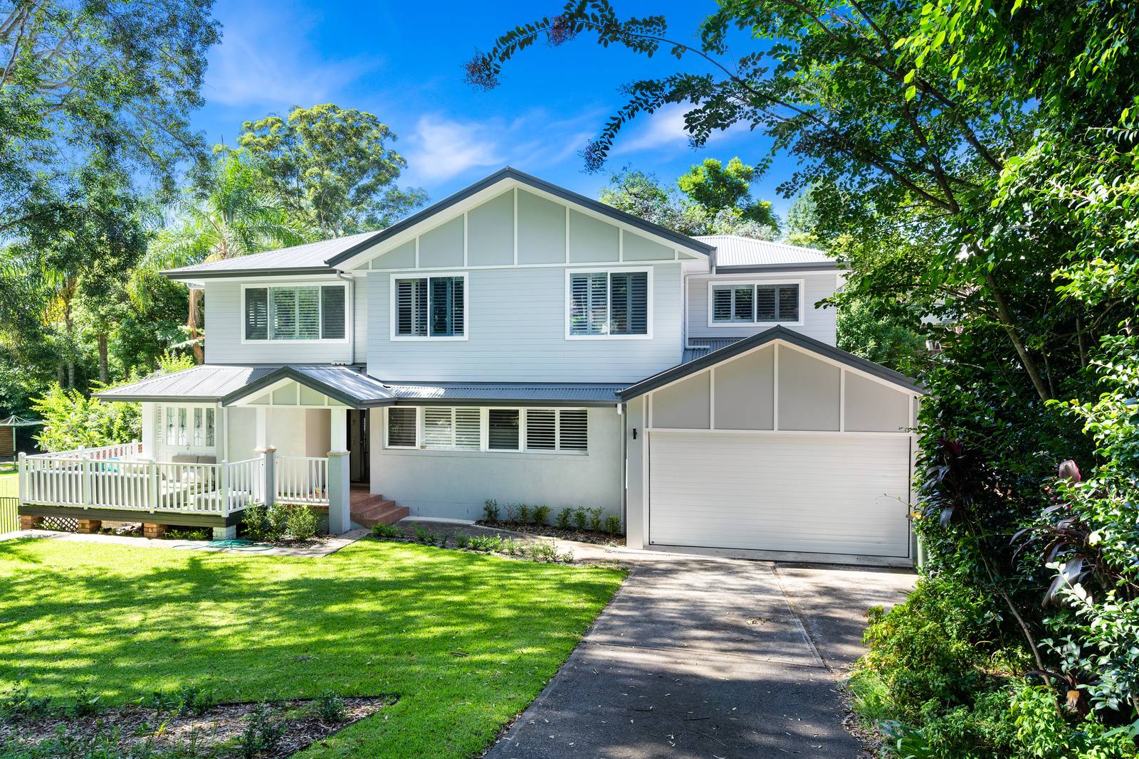 Two-story modern white house with large windows, a front porch, and an attached garage, surrounded by green trees, lush lawn, and landscaped garden on a sunny day.