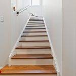 Second Storey Addition, Lane Cove A narrow, indoor staircase with wooden steps and white risers ascends between white walls, featuring a handrail on the left side and a window at the top landing letting in natural light.