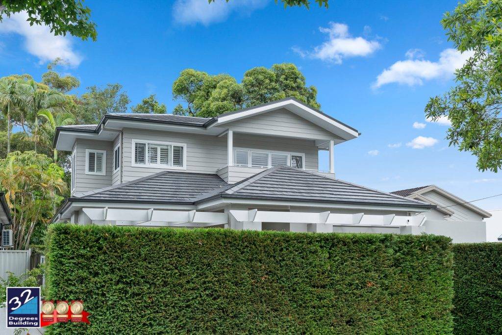 Second Storey Addition - Lane Cove --22 Modern two-story house with light grey siding and a dark tiled roof, surrounded by lush green trees and a tall, neatly trimmed hedge. A blue 32 Degrees Building logo is in the bottom left corner.
