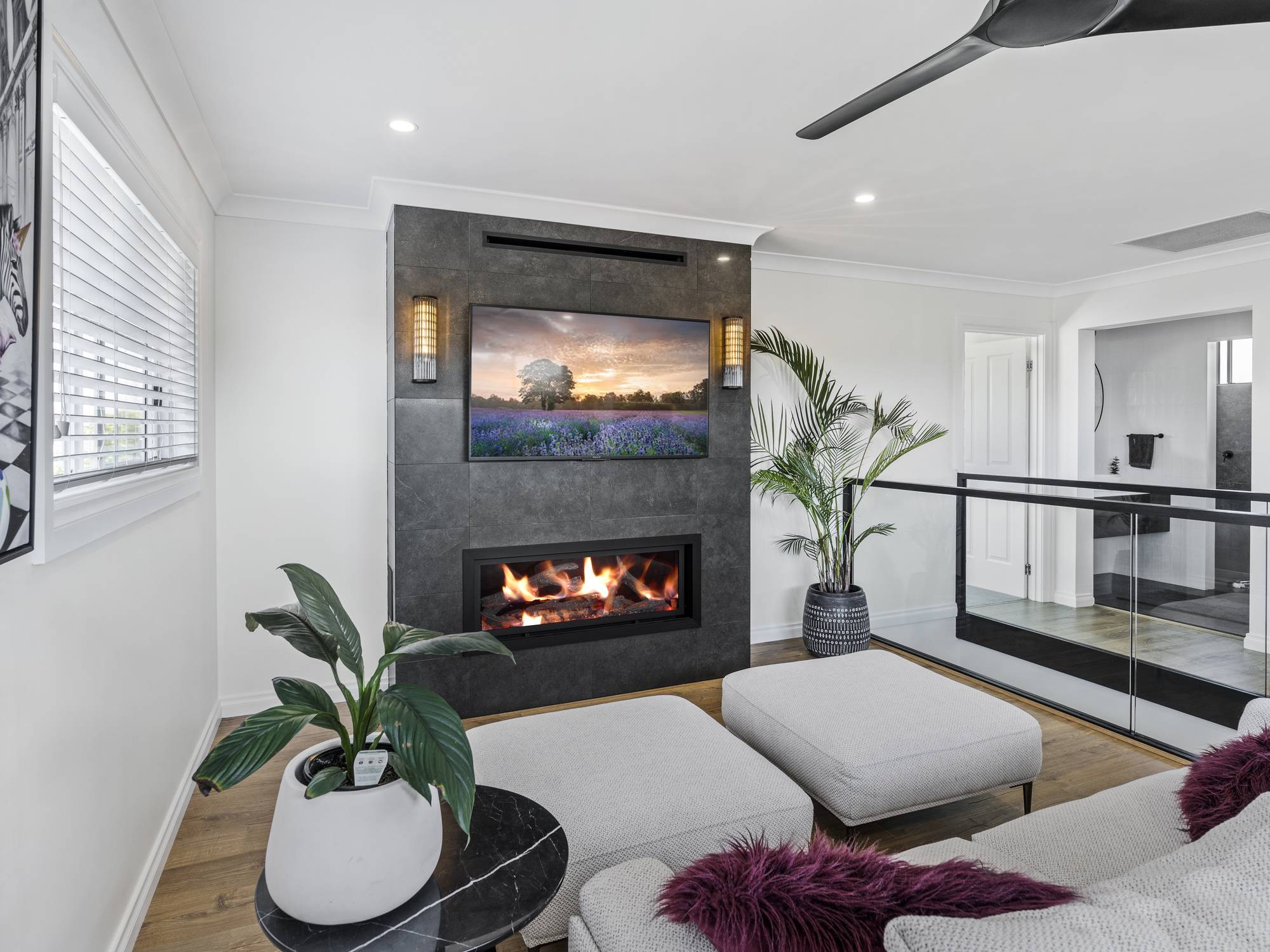 A modern living room with a fireplace and TV on a dark feature wall, light sofas, potted plants, wooden floor, and glass railing. Large windows let in natural light, and a ceiling fan is visible above.