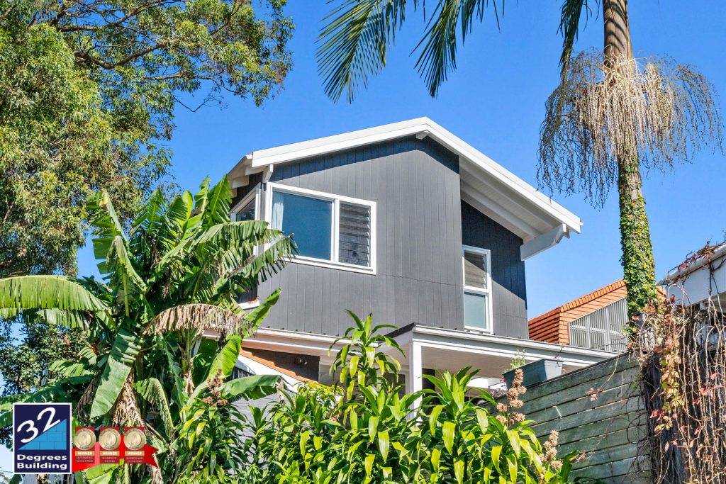 32 Degrees Building - Second Storey Addition Bronte (3 of 33) A modern two-story gray house with white trim surrounded by lush green tropical plants and palm trees. The sky is clear and blue. A 32 Degrees Building logo appears in the bottom left corner.