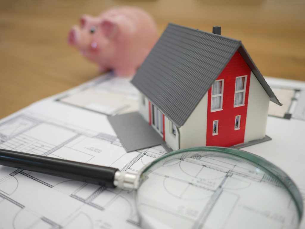 A model house sits on architectural blueprints next to a magnifying glass and a black pen, with a pink piggy bank blurred in the background.