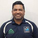 20230116_074722 A man with short dark hair and a beard smiles at the camera. He is wearing a navy blue collared shirt with white stripes and two logos, one green and one blue, on the front. The background is plain and light-colored.