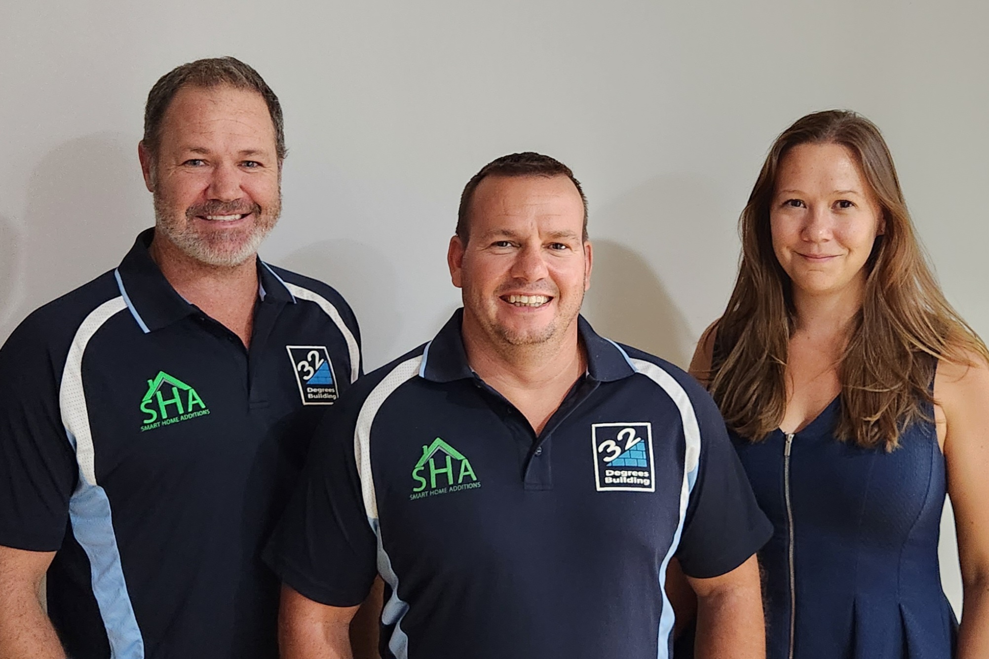 Denny, Alistair, Victoria Three people stand side by side smiling at the camera. Two men on the left wear matching dark shirts with company logos, while a woman on the right wears a sleeveless navy dress. The background is plain and light-colored.