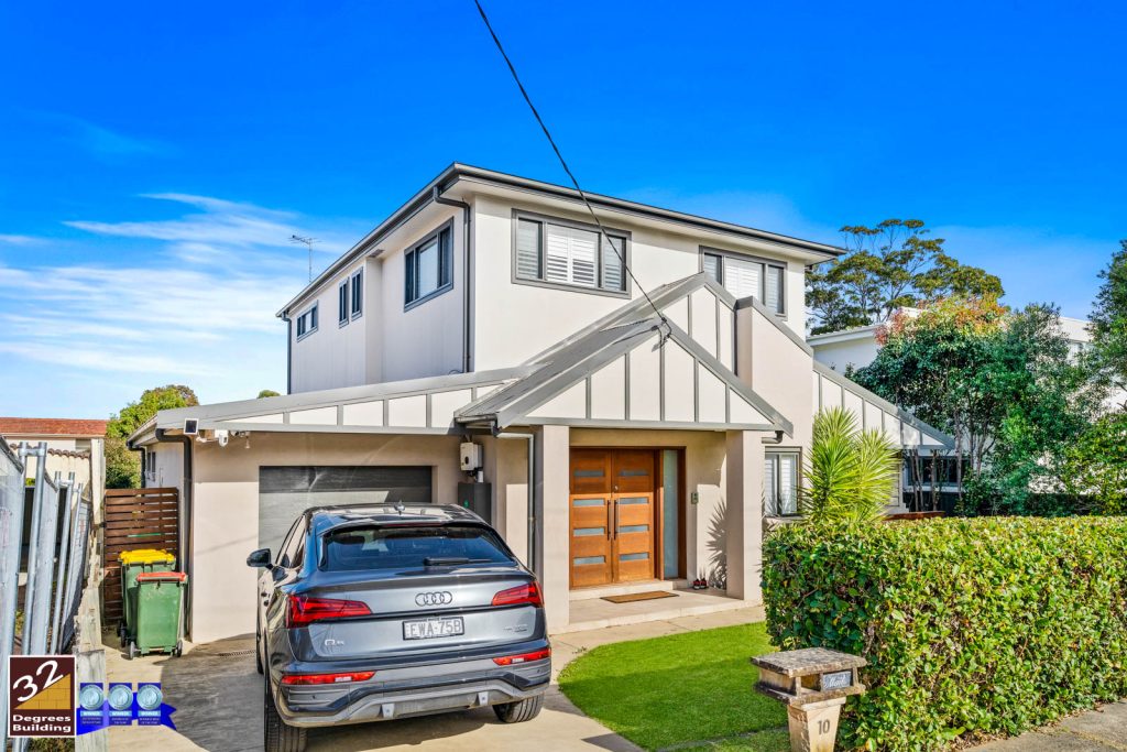 Second Storey Addition - Gladesville (2 of 16) Modern two-story house with light-colored exterior, wooden double front doors, and a silver Audi SUV parked in the driveway. Well-kept green hedges and trees surround the property under a clear blue sky.