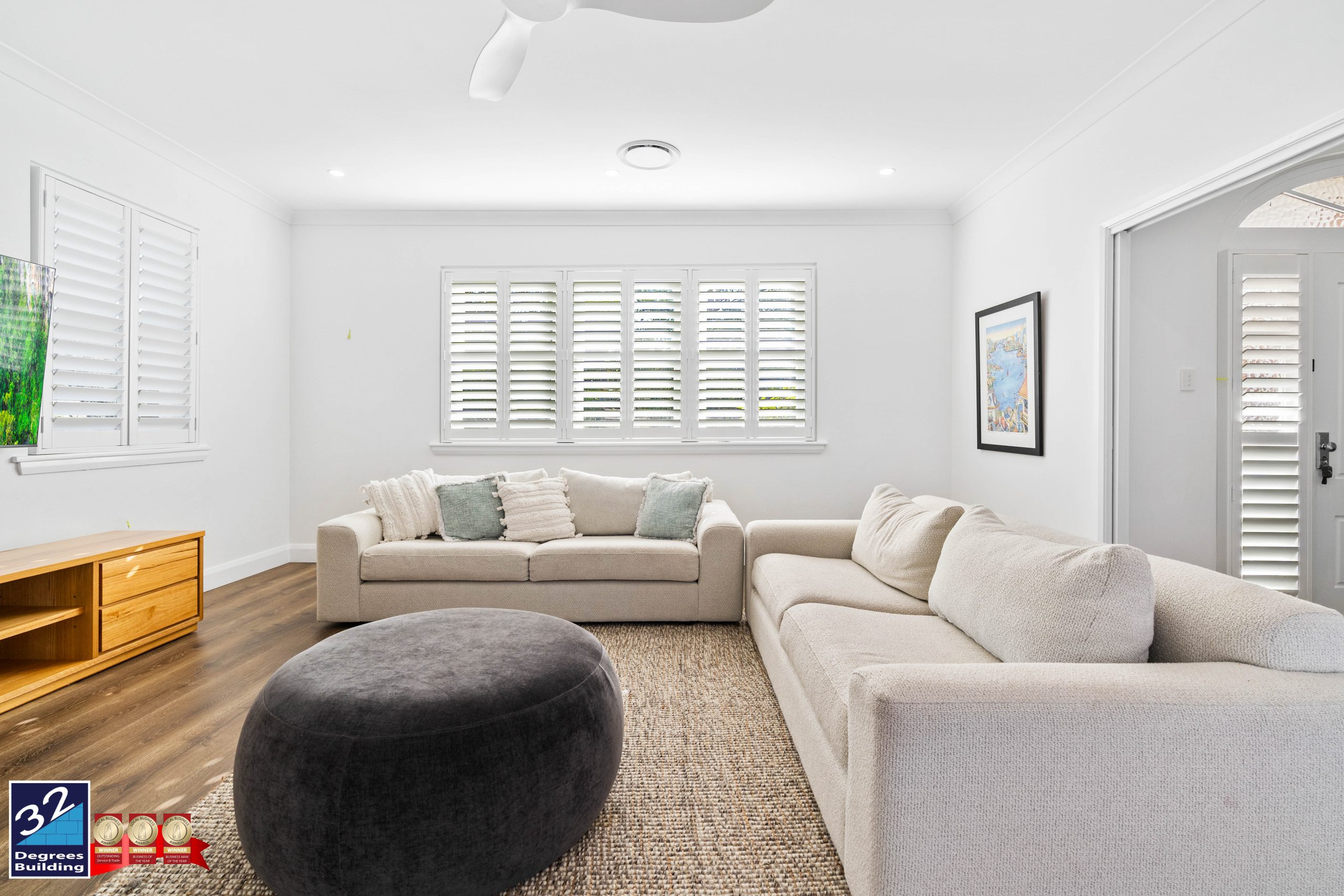 Second Storey Addition - Gladesville (9 of 89) A bright living room with two beige sofas, a large dark round ottoman, wood flooring, a TV, a light rug, and white shuttered windows letting in natural light. Artwork and a ceiling fan add to the modern decor.