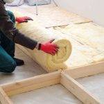 Insulation - Upgrade Between Floors A person wearing red gloves unrolls a sheet of yellow insulation material onto a wooden floor frame inside a room under construction or renovation.