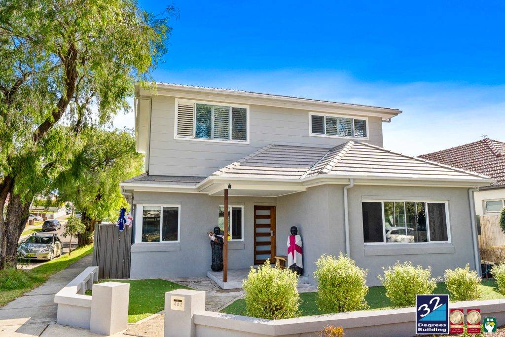 Second Storey Addition Kingsford (4 of 65) A modern two-story gray house with white trim, a small front porch, and well-kept landscaping. Decorative statues are by the entrance, and a 32 Degrees Building sign is in the front yard.