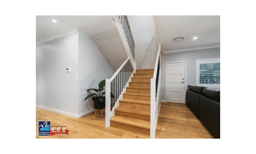 Staircase - Standard Inclusions Modern interior with a wooden staircase featuring white railings, light wood floors, a black sofa, a green potted plant, and a closed white door. Two award icons and a realty logo are visible in the lower left corner.