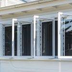Windows - Upgrade Casement Four white, side-hinged windows are open outward on a light-colored building, showing black window screens behind them. The roof eaves are visible above the windows.