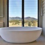 Windows - Upgrade Louvre A modern white freestanding bathtub is positioned in front of tall windows with a scenic view of hills, trees, and a clear sky outside.