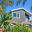 A modern two-story house with dark gray siding and large windows, surrounded by lush green tropical plants and trees under a clear blue sky. The 32 Degrees Building logo is in the bottom left corner.