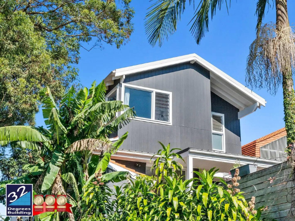 A modern two-story house with dark gray siding and large windows, surrounded by lush green tropical plants and trees under a clear blue sky. The 32 Degrees Building logo is in the bottom left corner.