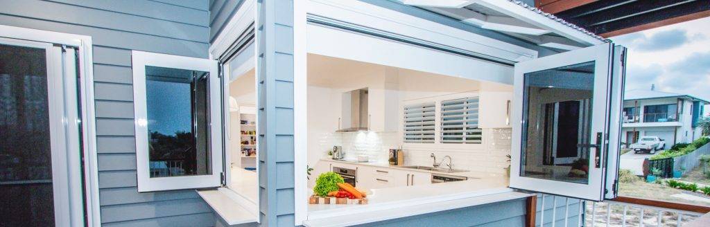A modern house with gray siding features large open windows connecting a bright kitchen to the outdoors; fresh vegetables sit on the white countertop by the window.