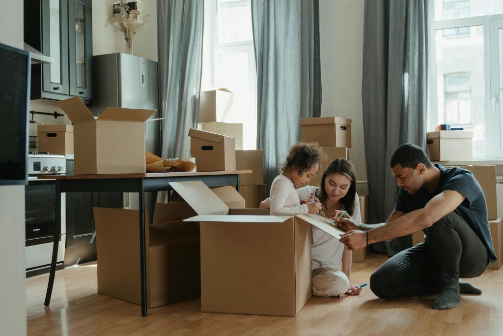 A family of three, including a young child, sits on the floor among cardboard boxes in a bright room, appearing to pack or unpack items together during a move.
