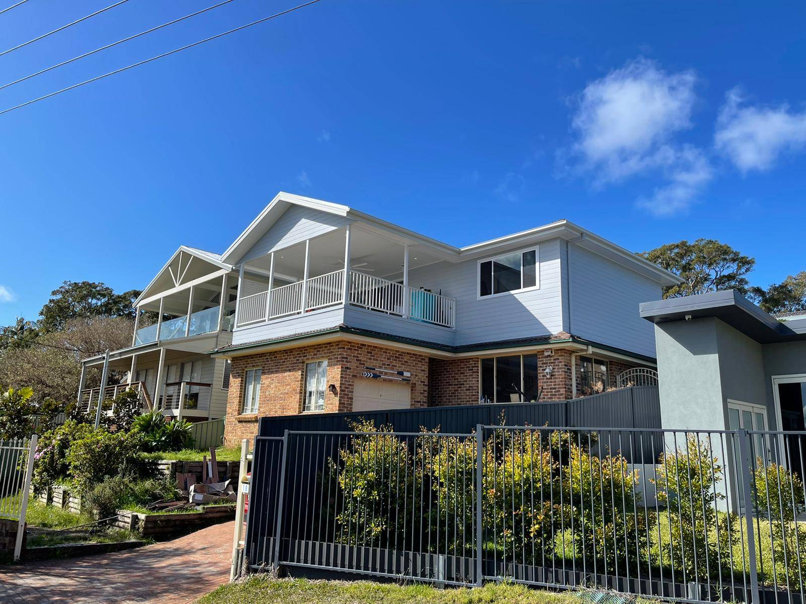 Two modern, two-story houses with balconies and large windows stand side by side behind a metal fence. The sky is clear and blue, and bushes and trees surround the homes.