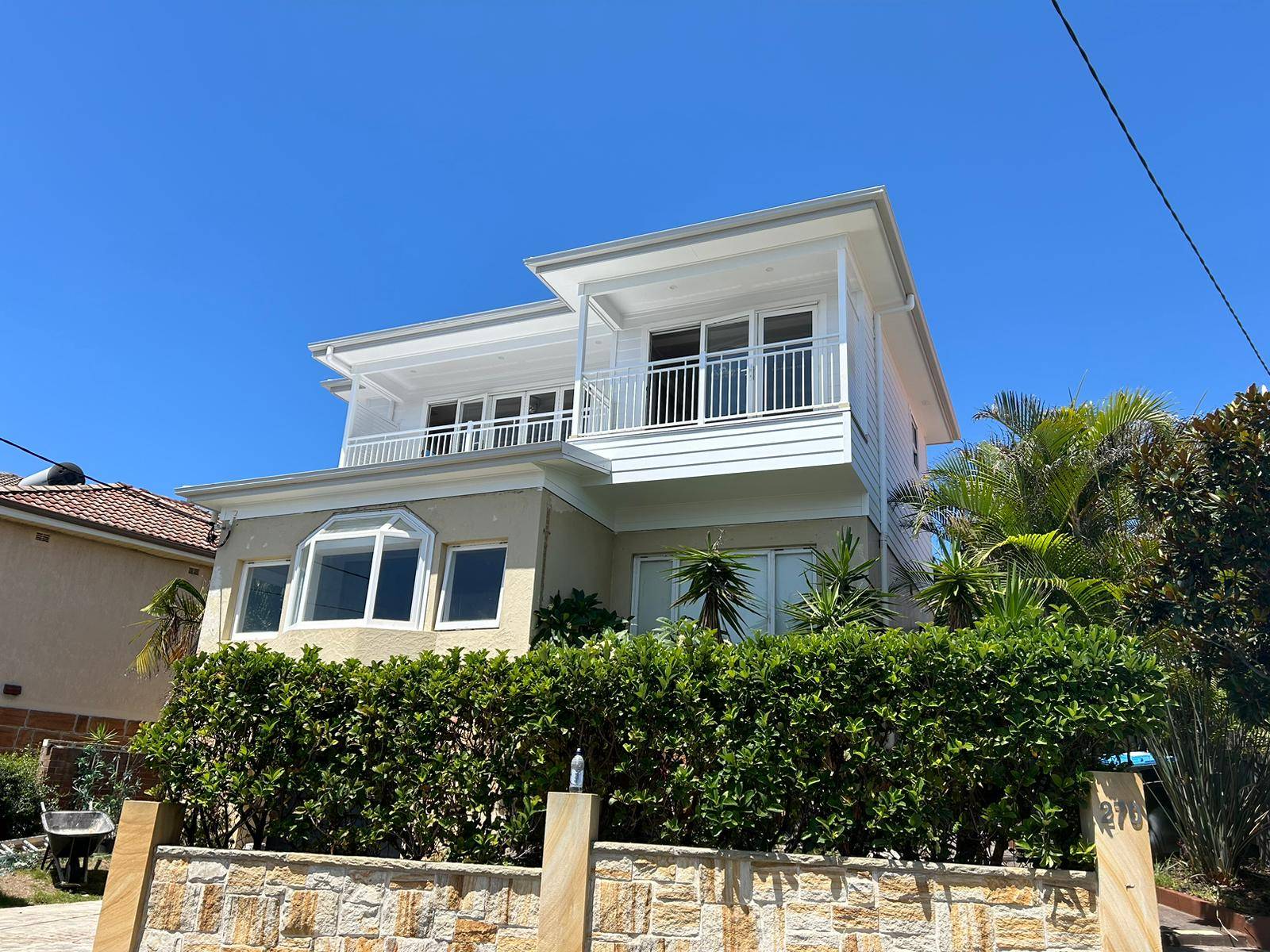 A two-story house with white upper walls and balconies, large windows, and a beige lower exterior, surrounded by greenery and a stone fence, under a clear blue sky.