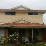 A two-story suburban house with a tiled red roof, cream upper walls, and brick lower walls, featuring a garage, front veranda with chairs, and a garden with blooming white roses.