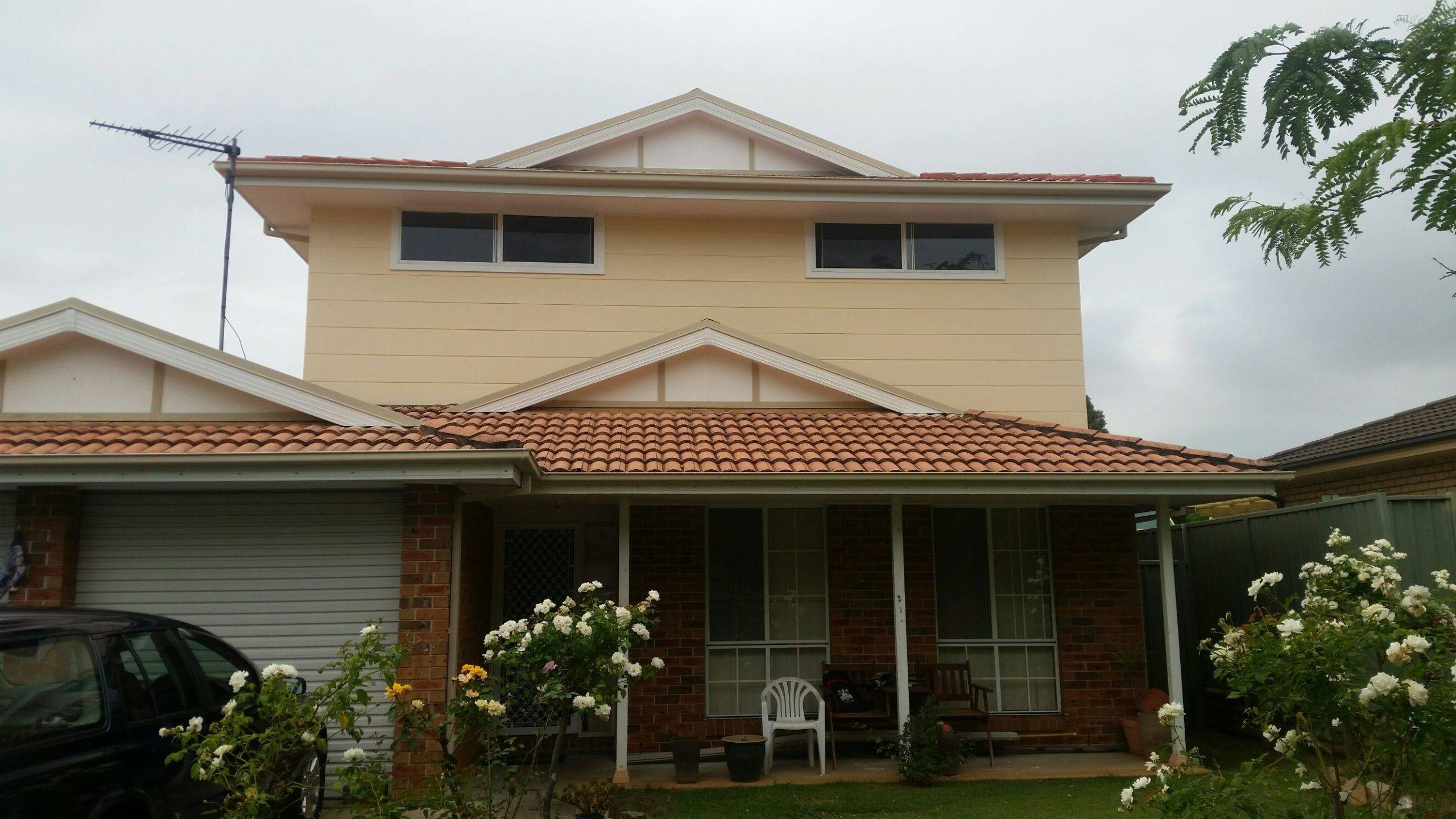 A two-story suburban house with a tiled red roof, cream upper walls, and brick lower walls, featuring a garage, front veranda with chairs, and a garden with blooming white roses.