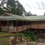 A wooden house with a green roof and wraparound porch sits on a sloped lot, surrounded by trees and rocks, with a gated driveway and patio furniture visible underneath the porch.