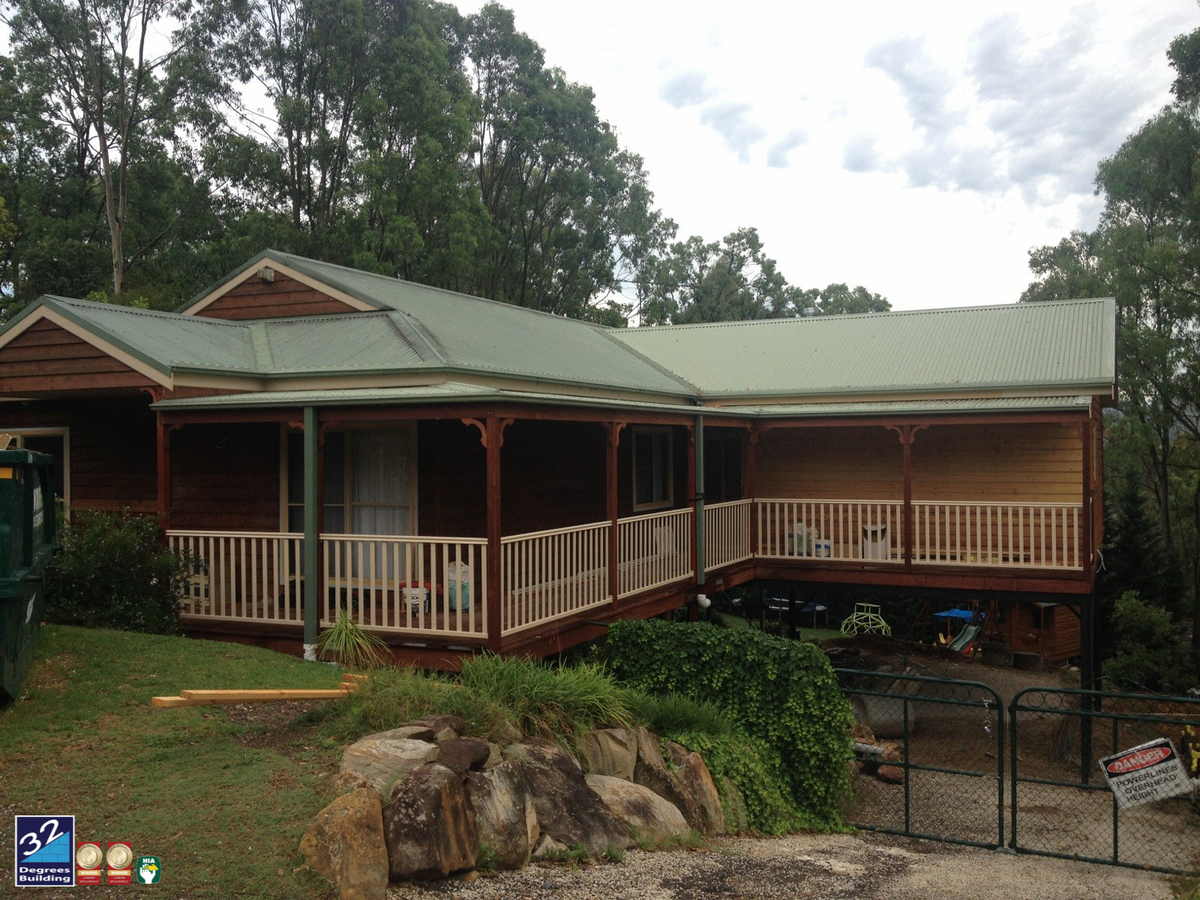 A wooden house with a green roof and wraparound porch sits on a sloped lot, surrounded by trees and rocks, with a gated driveway and patio furniture visible underneath the porch.