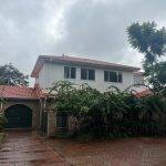A two-story house with a red-tiled roof, white upper walls, and brick lower walls. Lush greenery and vines cover the front, and a paved driveway leads to a closed garage. The sky is overcast and the ground is wet.