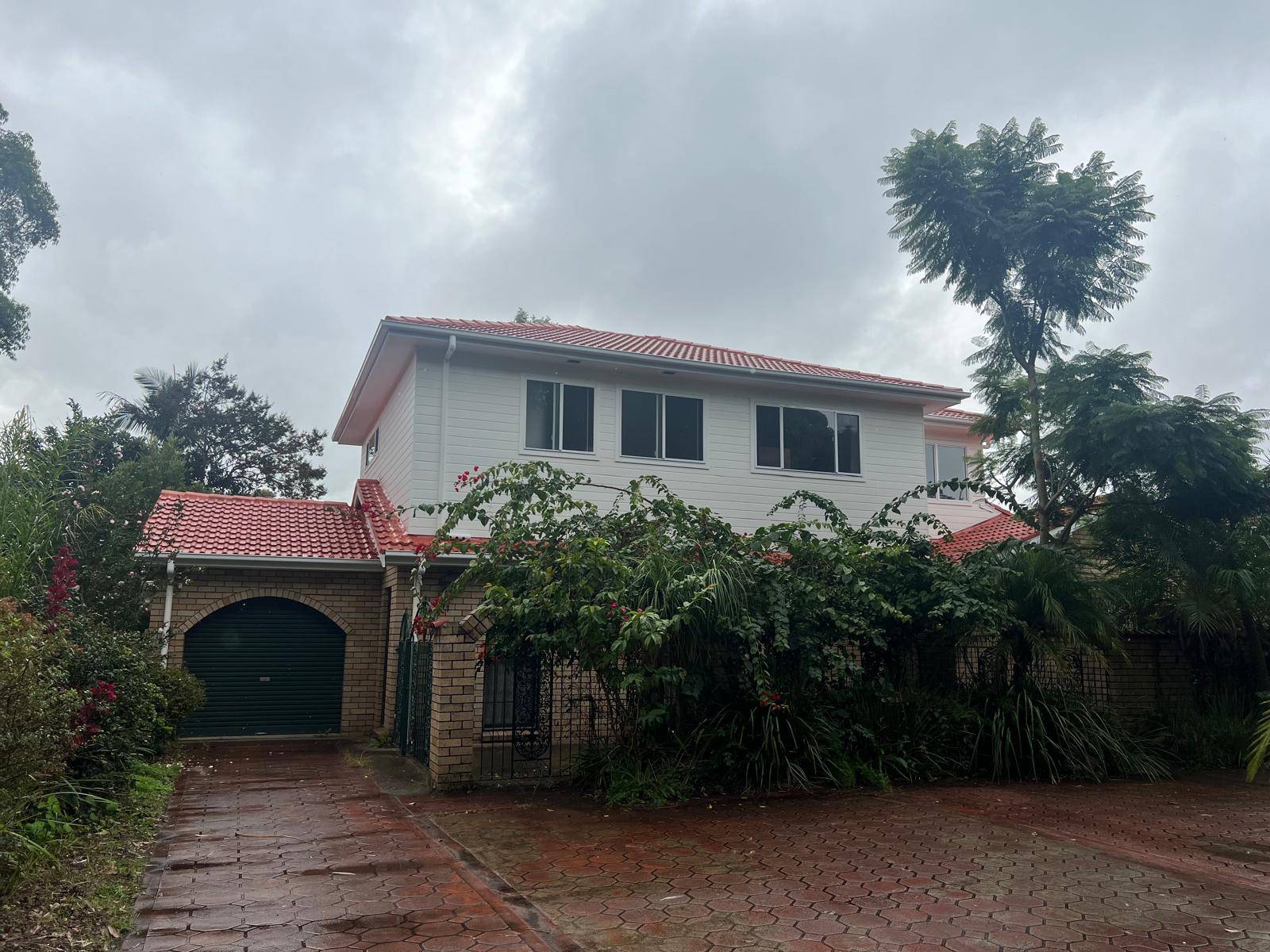 A two-story house with a red-tiled roof, white upper walls, and brick lower walls. Lush greenery and vines cover the front, and a paved driveway leads to a closed garage. The sky is overcast and the ground is wet.