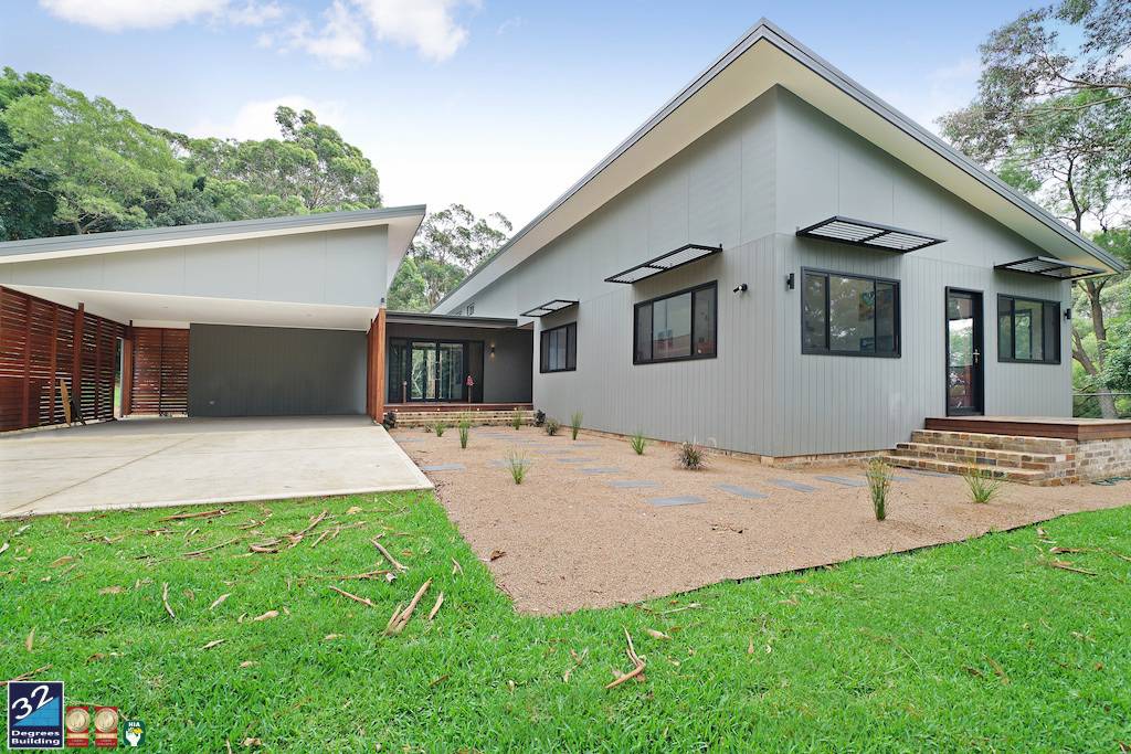 Modern single-story house with gray exterior, angular rooflines, large windows, and a covered carport. The front yard has a gravel pathway, young plants, and green grass, surrounded by trees under a partly cloudy sky.